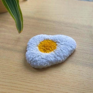 May include: A handmade, egg-shaped coaster with a white, fluffy exterior and a bright yellow center. The coaster is placed on a light-colored wooden surface, with a green plant visible in the background. The coaster is approximately 4 inches in diameter.