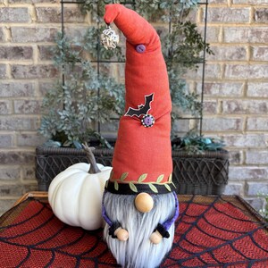 May include: A fabric gnome figurine with a tall, pointed orange hat featuring a small silver bell at the tip, a purple button, and a black bat applique. The gnome has a long gray faux fur beard, a round wooden nose, and a black band with green leaf patterns.