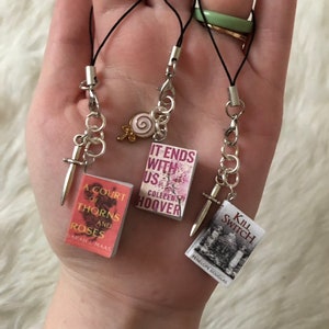 Peut inclure: Trois breloques miniatures en forme de livre avec des chaînes et des breloques argentées. Les breloques sont attachées à des cordons noirs. Les livres sont "A Court of Thorns and Roses" de Sarah J. Maas, "It Ends With Us" de Colleen Hoover et "Kill Switch" de Penelope Douglas.
