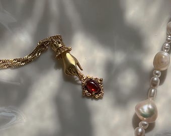 May include: A gold necklace with a pendant featuring a hand holding a red gemstone. The necklace is on a white surface with a pearl necklace beside it.
