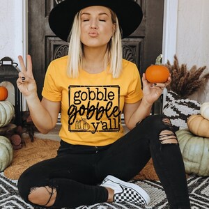 Può includere: Una donna indossa una maglietta gialla con il testo "gobble gobble y'all" in un riquadro nero. Tiene in mano una piccola zucca arancione.