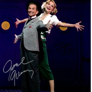 May include: A man and a woman performing on stage. The man is wearing a grey suit with a white shirt and a black hat. The woman is wearing a green skirt, a white top, and a white hat. The man has his arms outstretched, and the woman has her arms outstretched. The man is smiling, and the woman is smiling. The background is a dark blue stage.