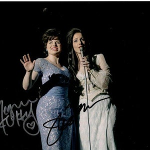 May include: Two women in vintage dresses singing into microphones on a dark stage. The woman on the left is wearing a blue lace dress and the woman on the right is wearing a white lace dress.