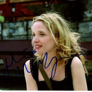 May include: A woman with long blonde hair is wearing a black sleeveless top with a brown strap. She is looking to the side and smiling. The background is blurred and out of focus.