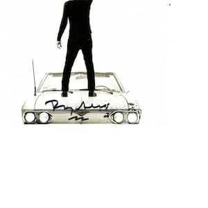 May include: Monochrome image of a person standing on a vintage car, holding a guitar overhead. The car's front features headlights and a signature. The person is wearing black clothing. The background is plain white.