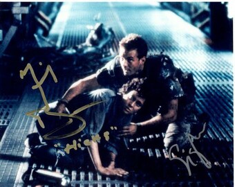 May include: A black and white photo of two people in a futuristic setting. The person on the left is lying on the ground, while the person on the right is standing over them. The person on the right is wearing a dark jacket and pants, while the person on the left is wearing a light shirt and pants. The person on the right has their hand on the person on the left's shoulder. The photo is signed by both people, with the person on the right's signature being "Sigourney Weaver" and the person on the left's signature being "Michael Biehn".