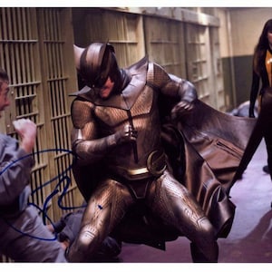 May include: A masked superhero in a brown and gold costume fights a man in a prison setting. The superhero is wearing a mask with a large, white owl face. A woman in a black and yellow costume stands in the background.