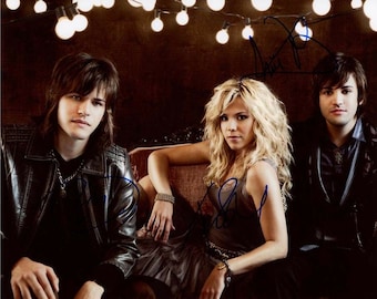 May include: A group of three musicians posing for a photo. The musicians are wearing casual clothing and are standing in front of a dark background with string lights. The woman in the middle is wearing a brown tank top and has long blonde hair. The man on the left is wearing a black leather jacket and has long dark hair. The man on the right is wearing a black button-down shirt and has short dark hair.
