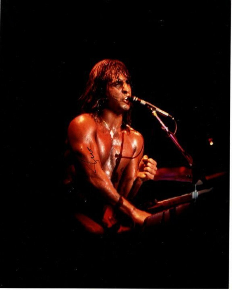 May include: A shirtless man with long brown hair sings into a microphone. He is wearing a microphone pack and a black strap around his waist. The background is dark.
