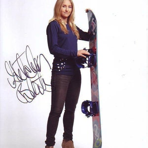 Puede incluir: Una mujer con una camisa azul de manga larga y pantalones vaqueros oscuros sostiene una tabla de snowboard con un diseño azul, rojo y blanco. Lleva botas marrones y tiene el pelo largo y rubio. La tabla de snowboard tiene una fijación azul y blanca.