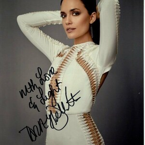 Può includere: Una donna in un abito bianco con un'applicazione di pizzo posa per una foto. Ha i capelli scuri e indossa orecchini. L'abito ha una scollatura profonda e una silhouette aderente. La donna ha firmato la foto con il messaggio "with love & light" e il suo nome.