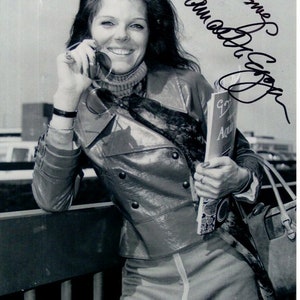 Puede incluir: Una fotografía en blanco y negro de una mujer con una chaqueta de cuero, una falda y un jersey de cuello alto. Lleva una revista y un bolso. La portada de la revista tiene un corazón y la palabra "Aqua". La mujer sonríe y mira a la cámara. La foto está firmada "Samantha Eggar".