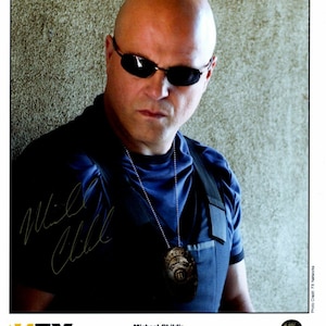 May include: A headshot of Michael Chiklis, who plays Detective Vic Mackey on the FX show "The Shield". He is wearing a blue shirt with a badge and a necklace. He is also wearing sunglasses and has a shaved head.