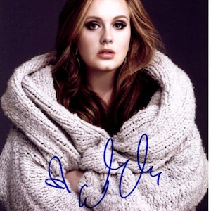 Puede incluir: Primer plano de una mujer con el pelo largo, ondulado y castaño rojizo que lleva un jersey de punto blanco. El jersey está drapeado sobre sus hombros y ella mira directamente a la cámara. La firma de la mujer es visible en tinta azul en el jersey.