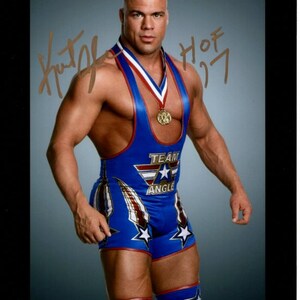 Puede incluir: Un luchador vistiendo un traje de lucha azul y rojo con estrellas y las palabras "Team Angle" en la parte delantera. El luchador tiene una medalla de oro alrededor del cuello y está firmando su nombre "Kurt Angle" con un marcador dorado.