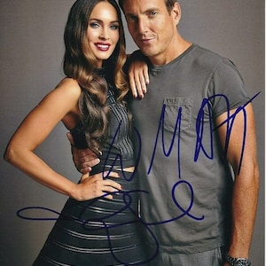 May include: A color photo of two people, a woman in a black dress and a man in a gray t-shirt, posing for a photo. The man is holding his arm around the woman's waist. The woman is smiling and looking at the camera. The man is smiling and looking at the camera. The man's signature is visible in the bottom right corner of the photo.