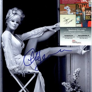 May include: Black and white photo of a woman sitting on a chair with her leg raised. She is wearing a white dress and has her hair styled in a bouffant. The woman is signing her name "Tifani Thiessen" in blue ink. The photo is authenticated by JSA.