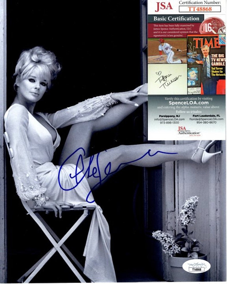 May include: Black and white photo of a woman sitting on a chair with her leg raised. She is wearing a white dress and has her hair styled in a bouffant. The woman is signing her name "Tifani Thiessen" in blue ink. The photo is authenticated by JSA.