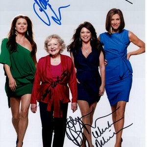 May include: Four women, including Betty White, pose for a photograph. They are all wearing different coloured dresses. Betty White is wearing a red jacket and black trousers. The women are all smiling and looking at the camera. The photograph is signed by all four women.
