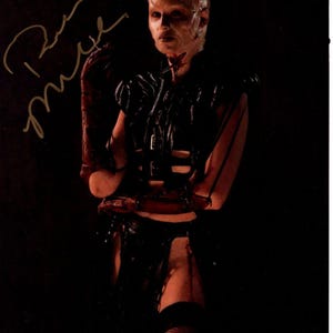 Könnte beinhalten: Signiertes Foto einer Figur aus einem Horrorfilm. Die Figur trägt ein schwarzes Lederoutfit mit detailliertem Design. Die Figur hat blasse Haut und einen rasierten Kopf. Die Signatur befindet sich in der oberen linken Ecke.