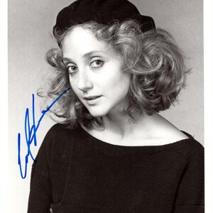 May include: Monochrome portrait of a woman in a black beret and dark sweater. The image is signed in blue ink. The woman has curly hair and is looking towards the camera.