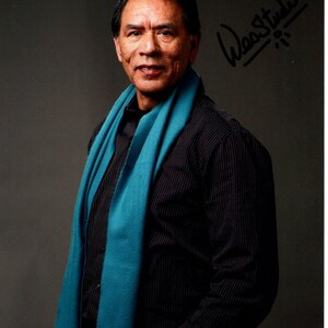 May include: A portrait of a man wearing a black pinstriped shirt and a teal scarf. The man is smiling and looking at the camera. The image has a signature in the upper right corner.
