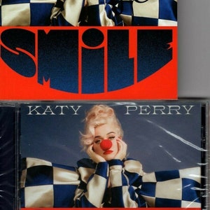 Puede incluir: Una caja de CD con un patrón de cuadros rojos y azules. La caja presenta una foto de Katy Perry con un disfraz de payaso con una nariz roja. El texto "KATY PERRY" y "SMILE" está impreso en la caja.