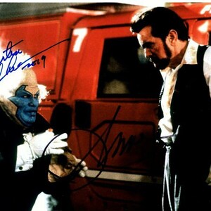 以下が含まれることがあります： 2人の男性の白黒写真。左側の男性は青と白の衣装を着ており、白いひげを生やしている。右側の男性は白いシャツの上に黒いベストを着ている。背景は、側面に7の数字が書かれた赤い消防車である。