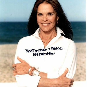 Może przedstawiać: Kobieta z długimi brązowymi włosami w białej koszuli i kilkoma bransoletkami na nadgarstku. Uśmiecha się i patrzy w obiektyw. Tło to plaża z niebieską wodą i białym piaskiem. Kobieta podpisała się "Best wishes & Peace,  Faye Dunaway" czarnym atramentem.