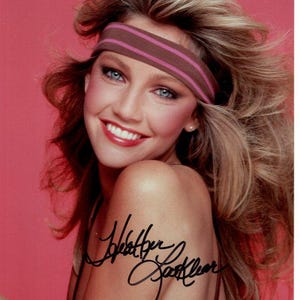 Puede incluir: Una mujer con el pelo largo y rubio que lleva una diadema marrón y rosa. Está sonriendo y mirando a la cámara. Su firma está escrita en tinta negra en su brazo. "Heather Locklear"