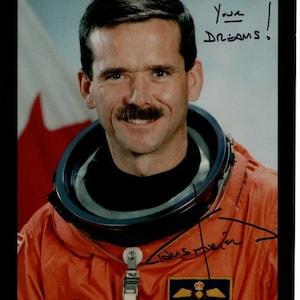 Puede incluir: Un hombre sonriente con un traje espacial naranja con una bandera canadiense de fondo. Lleva un cuello de lana negro y tiene bigote. El texto "Pursue Your Dreams!" está escrito en tinta negra sobre su cabeza. La firma del hombre está escrita en tinta negra debajo de su cuello de lana. El texto "Chris Hadfield" está escrito en tinta negra debajo de la firma.
