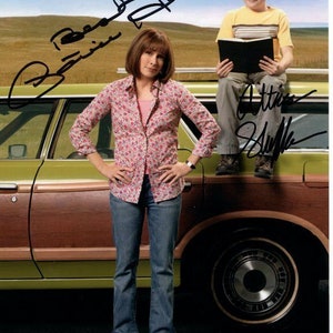 Puede incluir: Una mujer con una camisa de estampado rosa y blanco y pantalones vaqueros azules está de pie junto a una camioneta marrón y verde. Un niño pequeño con una camisa a rayas amarillas y pantalones vaqueros azules está sentado en la parte trasera de la camioneta sosteniendo un libro. La firma de la mujer es "Brenda Strong" y la del niño es "Atticus Shaffer".