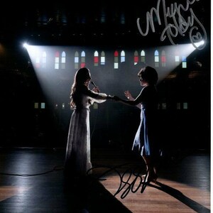May include: Two women in white and blue dresses stand on a stage with spotlights shining on them. They are holding hands and singing into microphones. The stage is in front of a brick wall with stained glass windows. The image is signed by the two women.