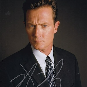 Op de afbeelding: Een man in een zwart pak met een wit overhemd en een gepatenteerde stropdas. Hij kijkt recht in de camera met een serieuze uitdrukking. Zijn handtekening is zichtbaar op het colbert.