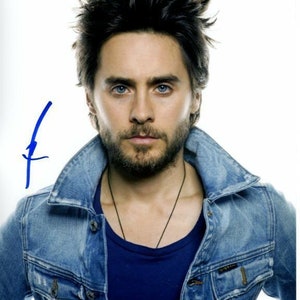 May include: A man with dark hair and a beard is wearing a blue denim jacket over a blue t-shirt. He is looking directly at the camera.