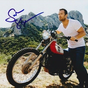 Puede incluir: Un hombre con una camiseta blanca y pantalones vaqueros azules está de pie junto a una motocicleta roja y negra. La motocicleta tiene un logotipo "Yamaha 750" en el lateral. El hombre está sujetando el manillar de la motocicleta. El fondo es un paisaje montañoso.