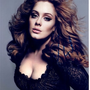 Puede incluir: Una mujer con el pelo largo y ondulado castaño, lleva un top de encaje negro. Está mirando a la cámara y tiene una expresión neutral. Su firma es visible en la imagen.