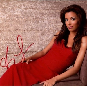 May include: A woman with long brown hair wearing a red sleeveless dress. She is sitting on a brown couch and smiling at the camera.