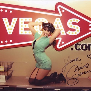 May include: A woman in a mint green romper poses in front of a red Vegas sign. The sign is shaped like an arrow with the word "VEGAS" in yellow letters. The woman is wearing black boots and stockings. The image is signed.