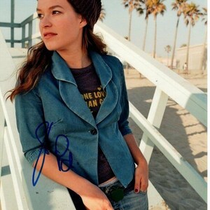 Könnte beinhalten: Eine Frau trägt eine blaue Jeansjacke über einem grauen T-Shirt mit dem Text "One Love Can Change The World" in Gelb. Sie trägt eine braune Mütze und eine blaue Jeans. Die Frau steht auf einem weißen Holzgeländer mit einem Strand im Hintergrund.