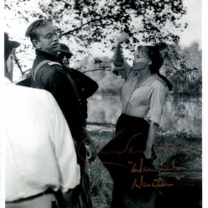 Peut inclure: Photographie en noir et blanc d'un plateau de tournage avec trois personnes. Une femme en chemise blanche et jupe foncée tient une bouteille et regarde vers la droite. Un homme en uniforme militaire regarde vers la gauche. La signature de la femme est visible dans le coin inférieur droit de l'image.