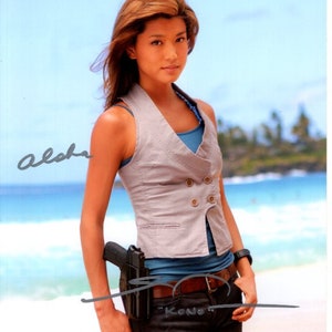 May include: A woman wearing a gray vest over a blue top and dark pants, standing on a beach. A black holster with a gun is attached to her belt. The word "Aloha" is written in cursive on the left side of the image, and "Kono" is written below the belt.