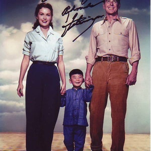 May include: A black and white photo of a family of three. A woman in a white shirt and dark pants holds the hand of a young boy in a blue shirt and pants. A man in a light brown shirt and brown pants holds the hand of the woman. The photo is signed "What a great memory - Rhonda Fleming"