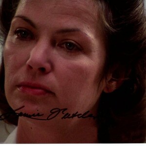 May include: A close-up portrait of a woman with brown hair and fair skin. The woman has a serious expression, with her eyes looking slightly downward. The image includes an autograph in the lower left corner.