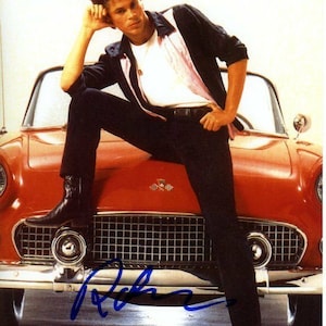 May include: A man in a black jacket and jeans poses on the hood of a red vintage convertible car. The car has a chrome grill and headlights. The man's signature is visible in blue ink on the front of the car.