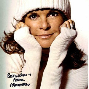 Może przedstawiać: Kobieta o brązowych włosach ma na sobie biały wełniany beret i pasujący sweter. Trzyma ręce przy twarzy i uśmiecha się. Obraz jest podpisany "Najlepsze życzenia i pokój,  [nierozpoznany podpis]".