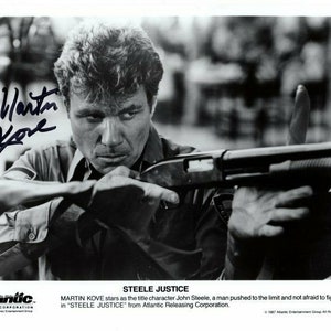 May include: Black and white photo of an actor holding a shotgun. The actor is looking directly at the camera. The photo is signed "Martin Kove". The text "STEELE JUSTICE" is printed at the bottom of the photo. The text "MARTIN KOVE stars as the title character John Steele, a man pushed to the limit and not afraid to fight back in "STEELE JUSTICE" from Atlantic Releasing Corporation." is printed below the title.