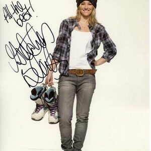 Puede incluir: Una mujer con un gorro negro, una camisa a cuadros, un top blanco, pantalones vaqueros grises y un cinturón marrón. Está sosteniendo un par de botas de esquí azules y blancas. El texto "All the best!" está escrito en tinta negra sobre un fondo blanco.