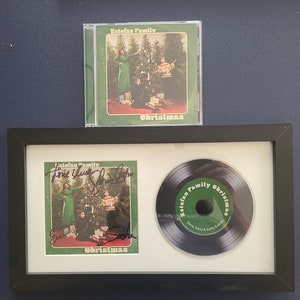 Puede incluir: Un álbum de Navidad autografiado de la familia Estefan, enmarcado. La portada del álbum presenta una foto de la familia frente a un árbol de Navidad. El álbum está firmado por Gloria, Emily y Lyle Estefan.