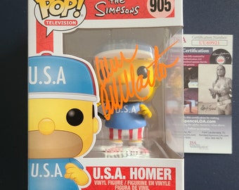 May include: A Funko Pop! Vinyl figure of Homer Simpson from The Simpsons TV show. Homer is wearing a blue and red outfit with a white baseball cap that says "U.S.A." on it. The figure is in a clear plastic box with a red and white background. The box has the Funko Pop! logo and the words "The Simpsons" and "905" on it. The figure is signed by the voice actor Dan Castellaneta.